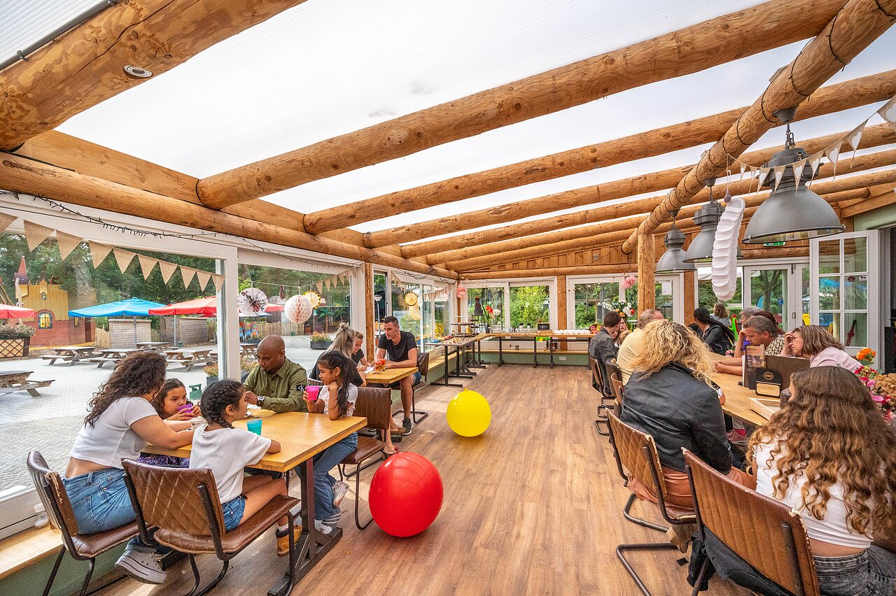 Familles et enfants d�nant au restaurant couvert avec vue sur l'aire de jeux au camping CAPFUN De Scheepsbel � Doornspijk.