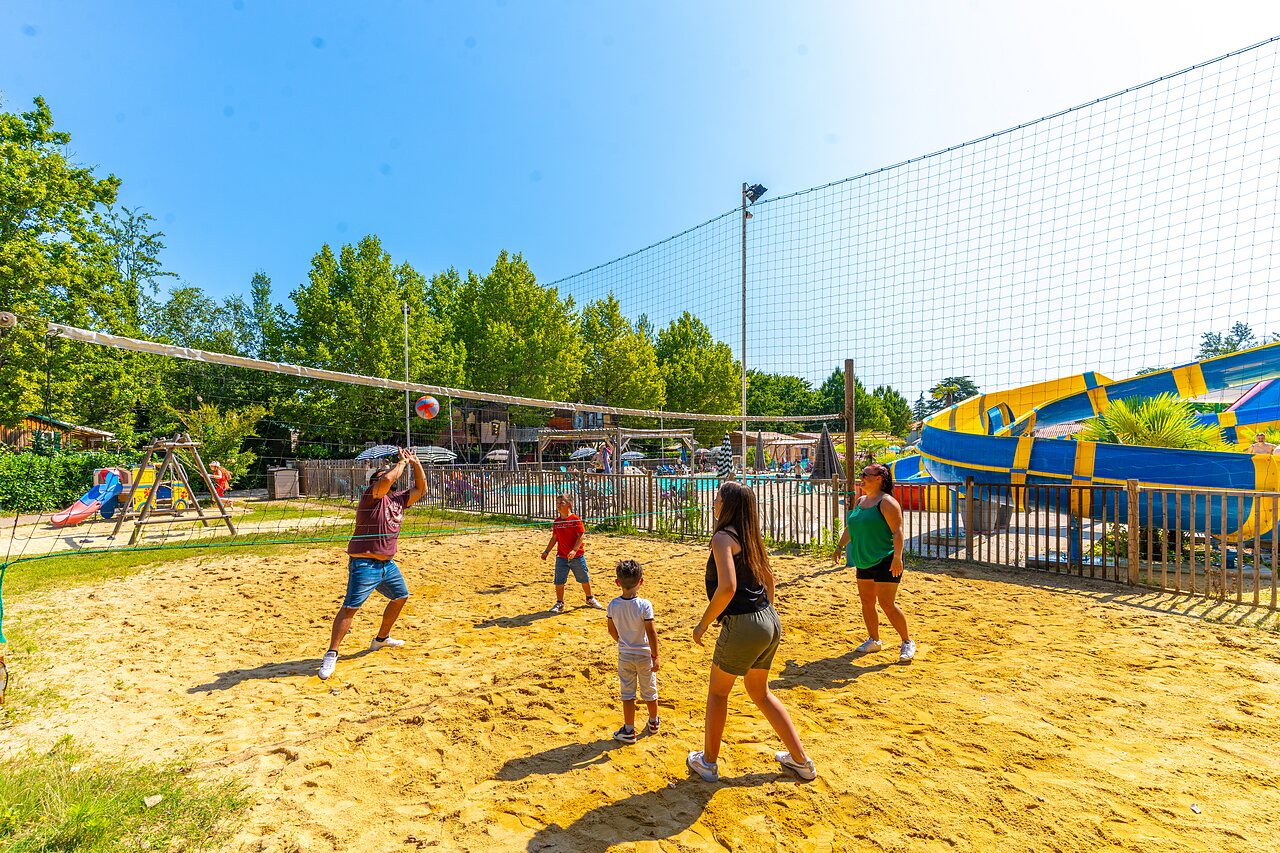 Famille jouant au beach-volley sur terrain de sable, toboggans aquatiques au camping CAPFUN Duravel.